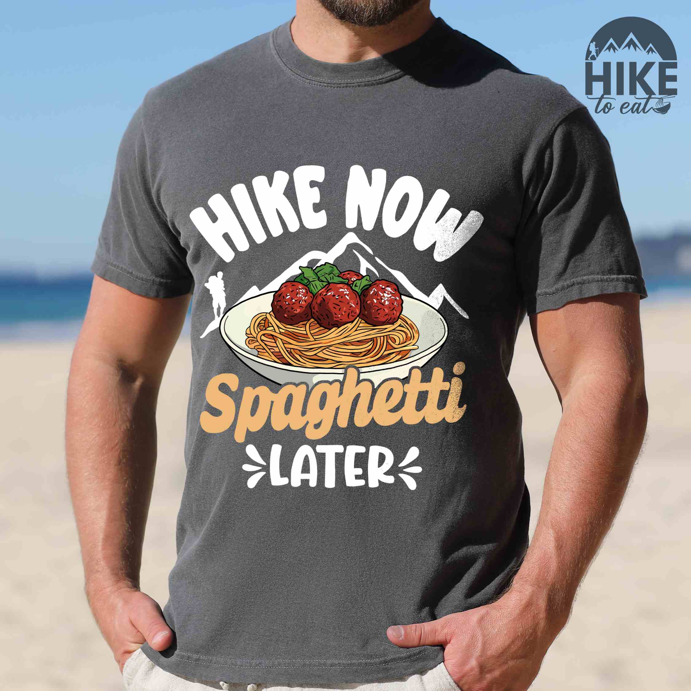 Man wearing pepper "Hike now spaghetti later" colored vintage tee with bold typography, mountains, birds, illustrated plate of spaghetti meatballs, and backpackers for pasta lovers.