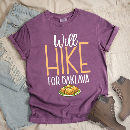Berry (Dark Violet) t-shirt with white and peach text reading "Will HIKE FOR BAKLAVA". "Will" appears in white script at the top, "HIKE" in large peach letters in the middle, and "FOR BAKLAVA" in white at the bottom. Features an illustration of baklava pieces with honey and pistachios on a green plate below the text. Features a classic crew neck and casual rolled sleeves, styled alongside light wash denim and brown hiking boots. 