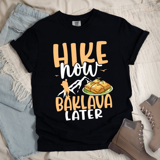 Black t-shirt with peach and white text reading "HIKE now BAKLAVA LATER". "HIKE" appears in large peach letters at the top, "now" in white script with small birds, and "BAKLAVA" in peach with "LATER" in white at the bottom. Features white mountain silhouettes, a peach hiker silhouette on the left, and an illustration of baklava pieces with pistachios on a green plate. Features a classic crew neck and casual rolled sleeves, styled alongside light wash denim and brown hiking boots. 