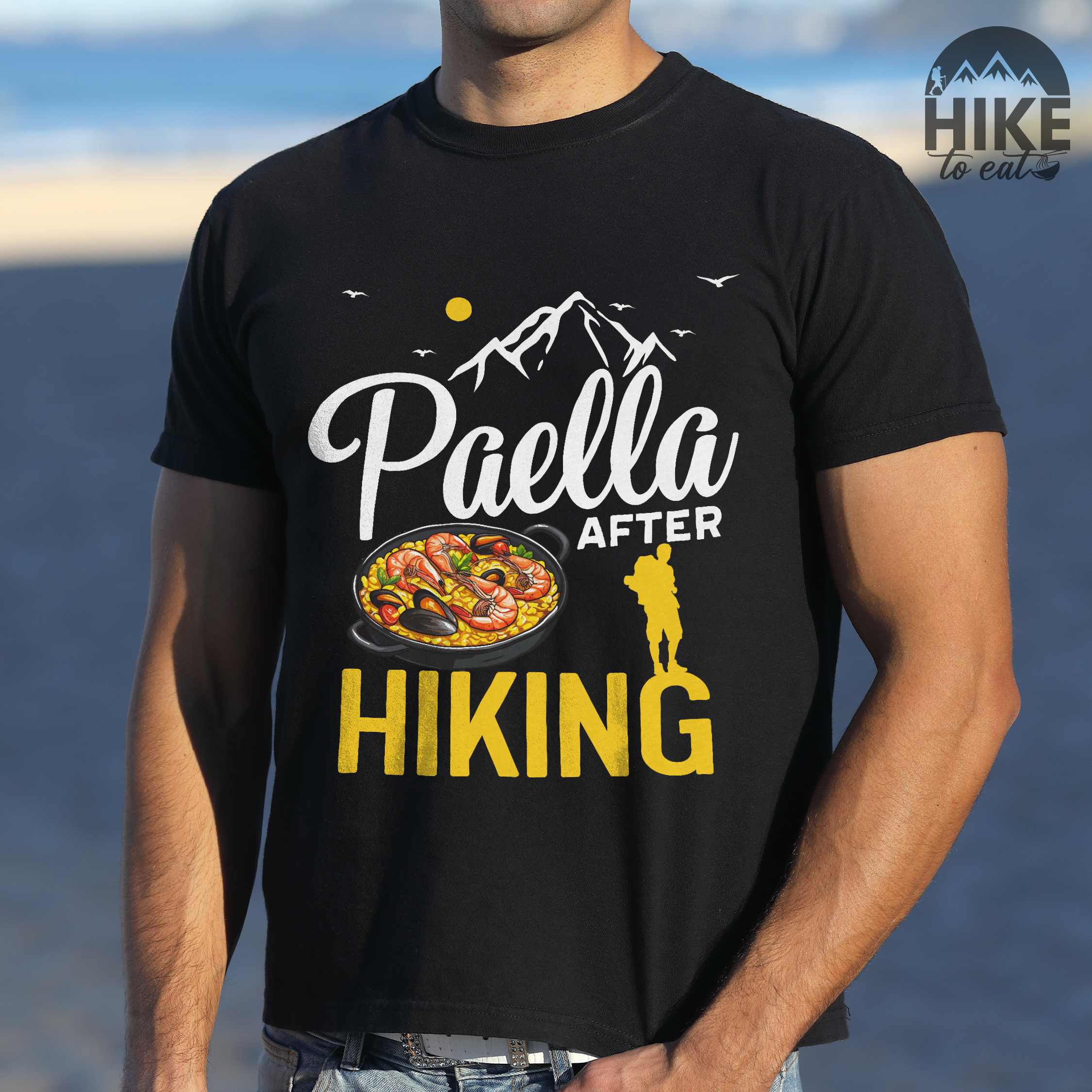 Man wearing black "Paella after hiking" colored vintage tee with bold typography, mountains, birds, illustrated paella platter, and backpackers for rice lovers.