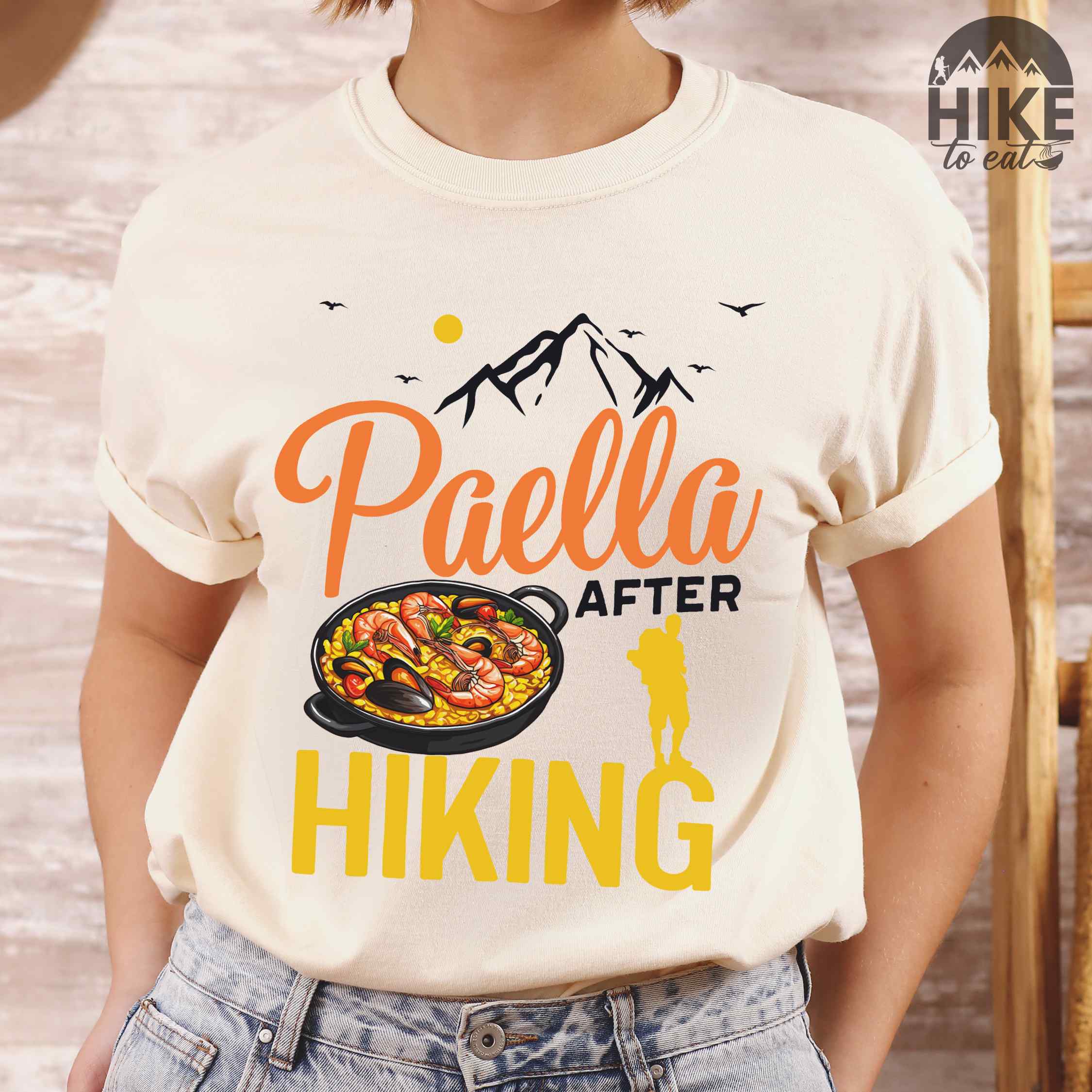 Woman wearing ivory "Paella after hiking" colored vintage tee with bold typography, mountains, birds, illustrated paella platter, and backpackers for rice lovers.