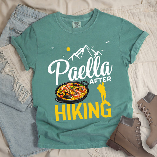 Light Green t-shirt with white and yellow text reading "Paella AFTER HIKING". "Paella" appears in white script at the top, "AFTER" in white text in the middle, and "HIKING" in large yellow letters at the bottom. Features white mountain silhouettes, birds, and a yellow sun. A yellow hiker silhouette appears between "AFTER" and on top of the letter "G" in "HIKING" on the right, with a detailed illustration of a paella dish with seafood in a black pan to the left of the hiker.
