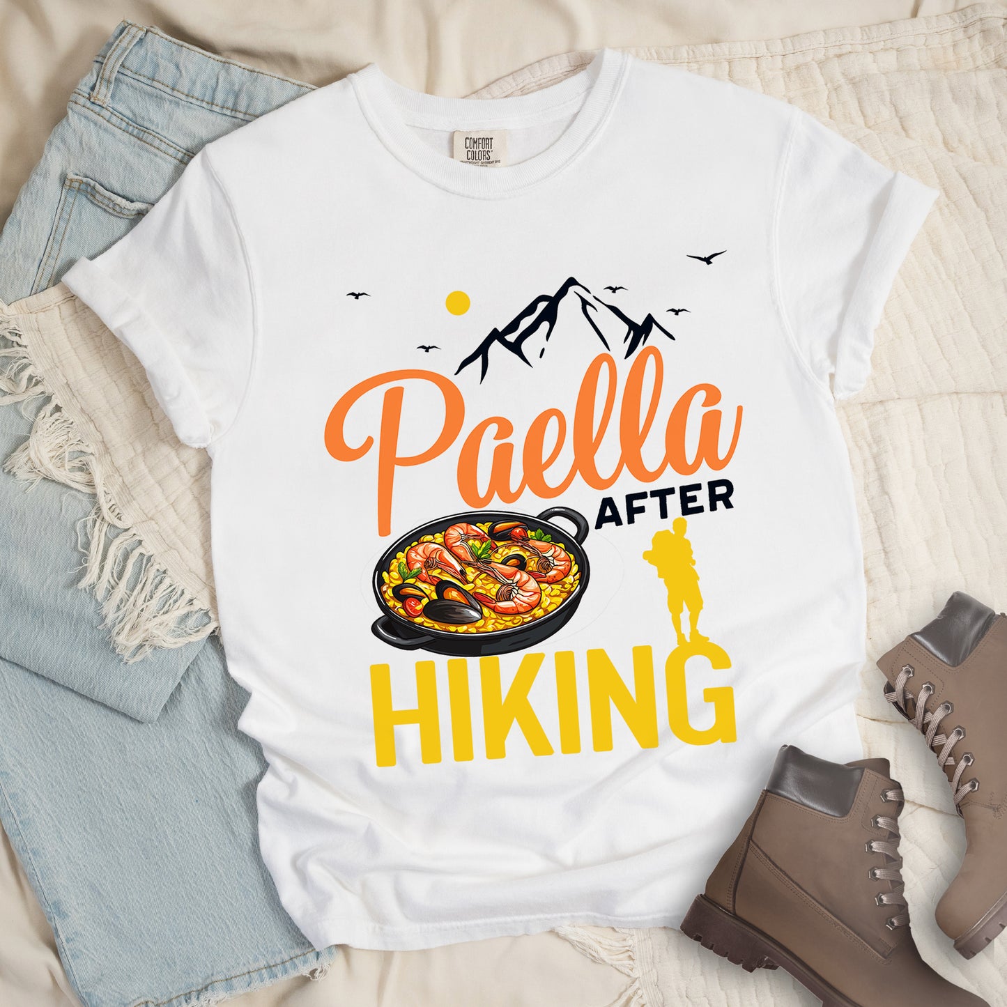 White t-shirt with black, orange and yellow text reading "Paella AFTER HIKING". "Paella" appears in orange script at the top, "AFTER" in black text in the middle, and "HIKING" in large yellow letters at the bottom. Features black mountain silhouettes, birds, and a yellow sun. A yellow hiker silhouette appears between "AFTER" and on top of the letter "G" in "HIKING" on the right, with a detailed illustration of a paella dish with seafood in a black pan to the left of the hiker.