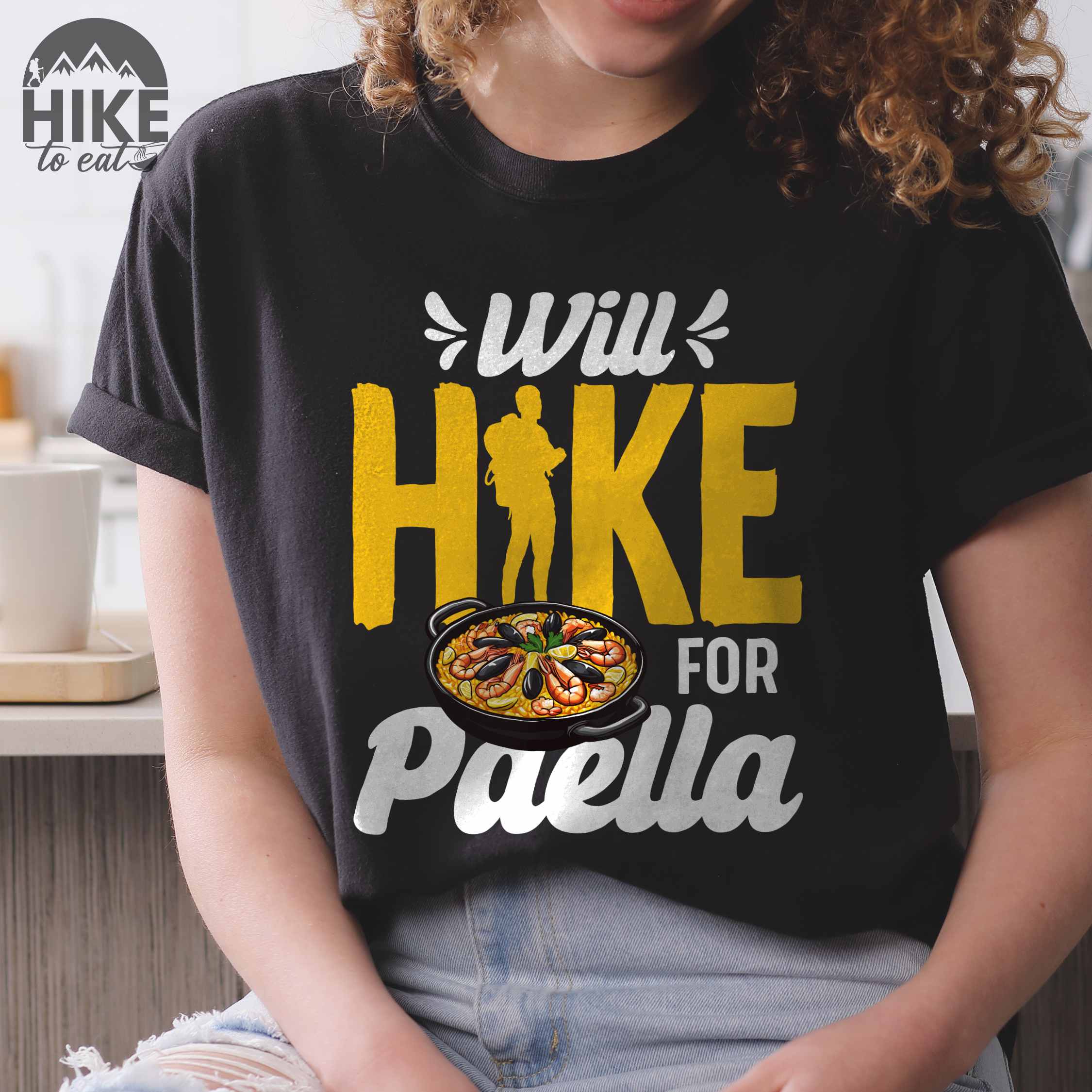Woman wearing black "Will Hike for Paella" colored vintage tee with bold typography, illustrated paella platter, and backpackers for rice lovers.