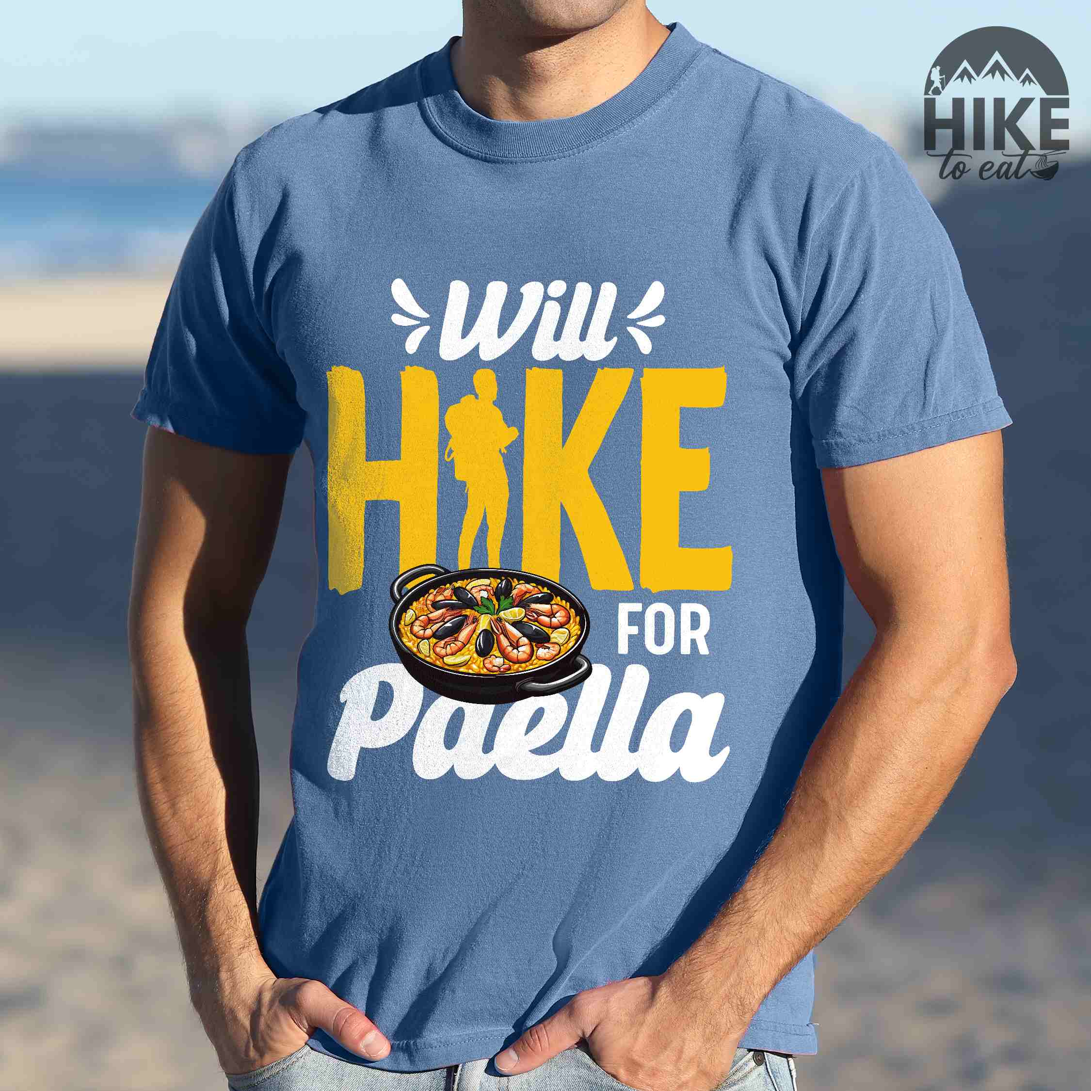 Man wearing blue jean "Will Hike for Paella" colored vintage tee with bold typography, illustrated paella platter, and backpackers for rice lovers.