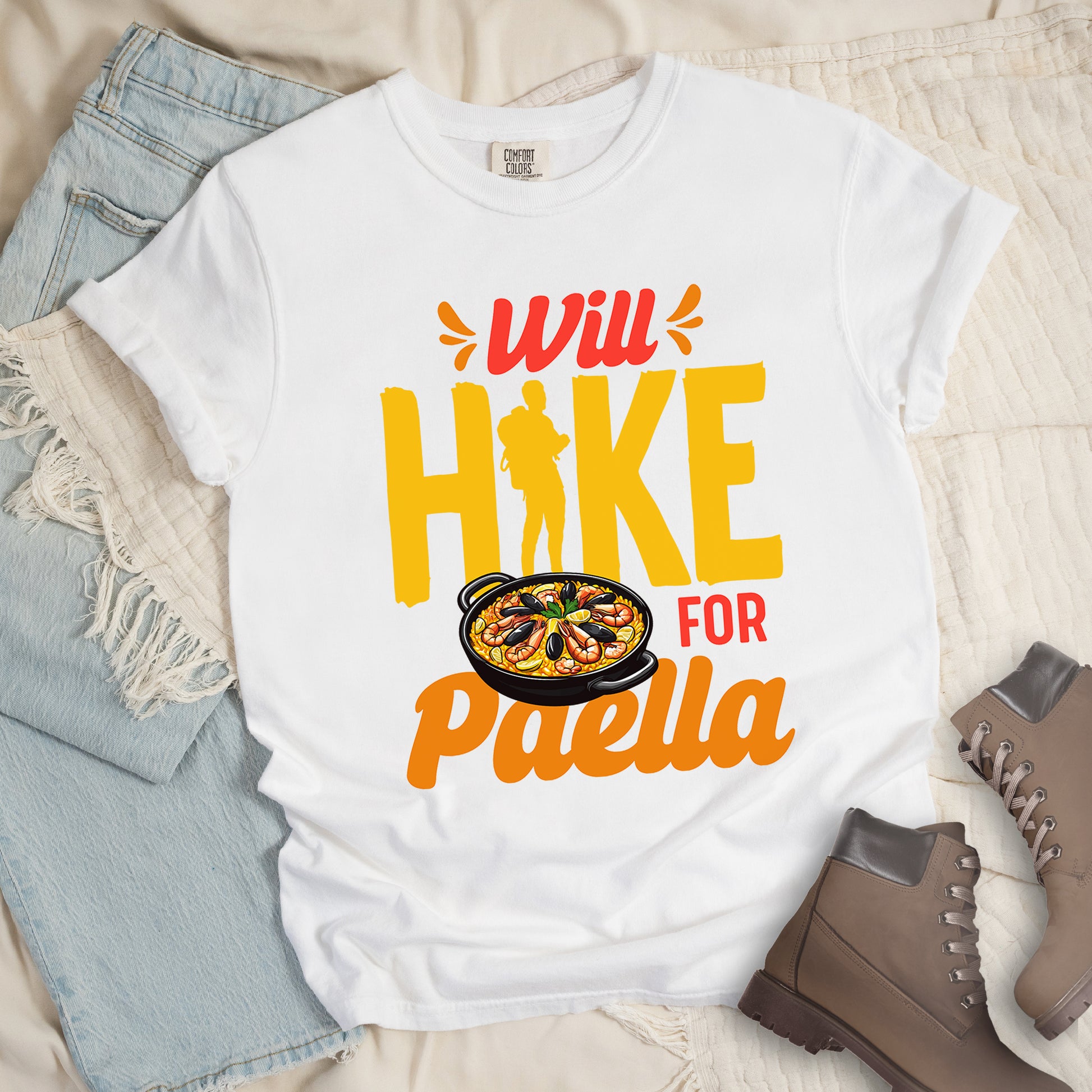 White t-shirt with red, orange and yellow text reading "Will HIKE FOR Paella". "Will" appears in red script at the top with small decorative accents, "HIKE" in large yellow letters with a yellow hiker silhouette integrated into the letter "I", "FOR" in red, and "Paella" in orange script at the bottom. Features an illustration of a paella dish with seafood in a black pan to the left of "FOR" and above "Paella".