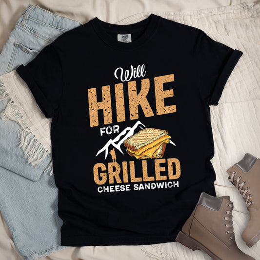 Black t-shirt with text  "Will HIKE FOR GRILLED CHEESE SANDWICH". "Will" appears in white script at the top, "HIKE" in large bold distressed golden-brown block letters, "FOR" in white, "GRILLED" in bold distressed golden-brown block letters, and "CHEESE SANDWICH" in white at the bottom. Features white mountain silhouettes, a golden-brown hiker silhouette, and an illustration of a grilled cheese sandwich with toasted bread and yellow cheese inside. 