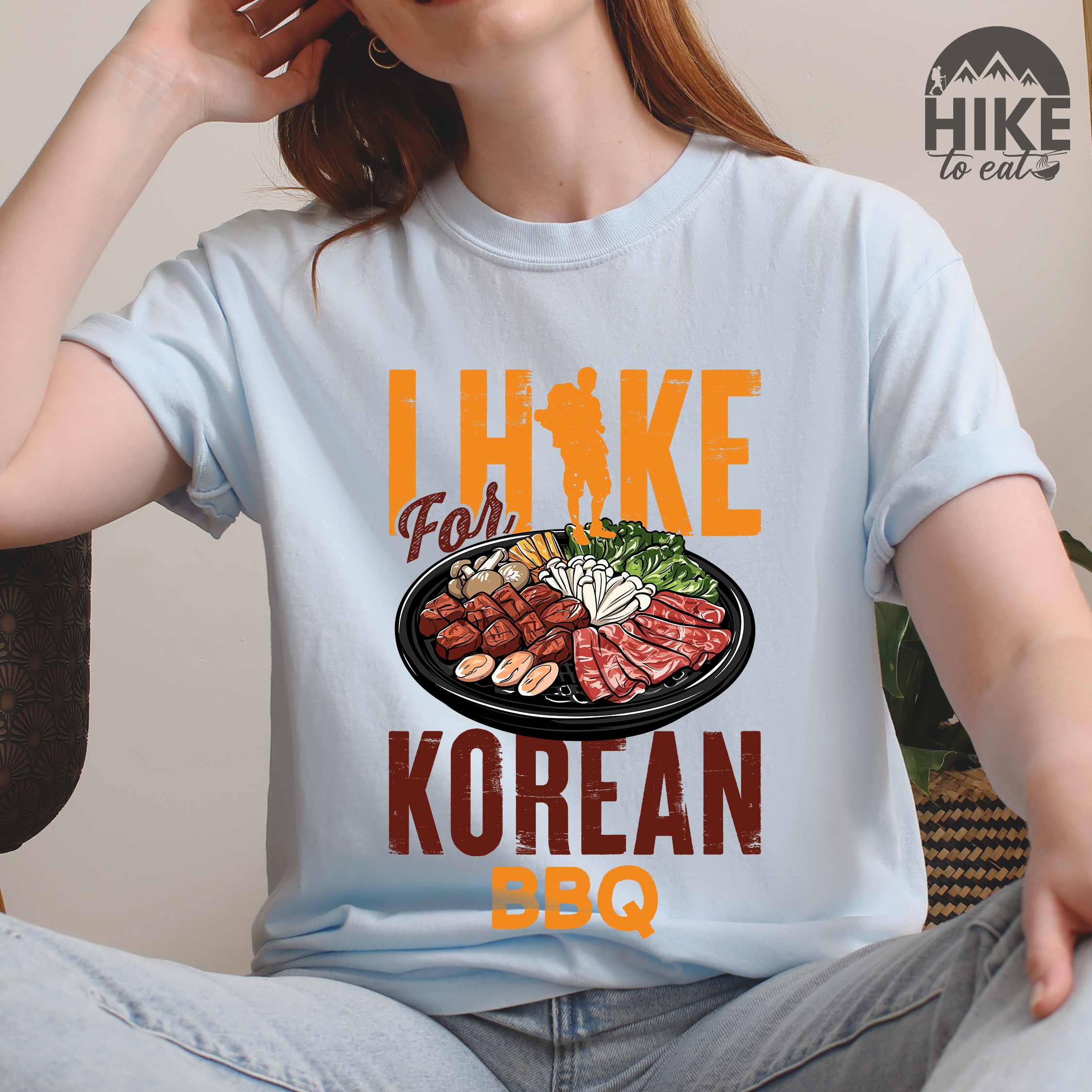 woman wearing chambray "I hike for korean bbq" colored vintage tee with bold typography, illustrated grilling platter korean bbq, mountains, birds, and backpacker for kbbq lovers.
