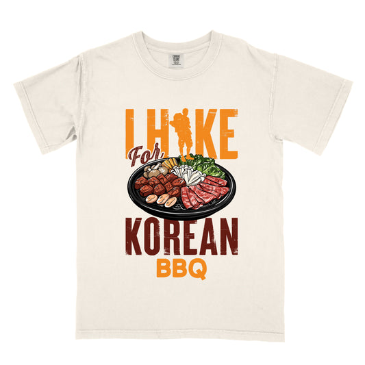 Ivory (Cream White) t-shirt with orange and red text reading "I HIKE For KOREAN BBQ". "I HIKE" appears in large distressed orange letters at the top with a hiker silhouette integrated into the letter "I", "For" in red script in the middle, "KOREAN" in large distressed red letters, and "BBQ" in orange at the bottom. Features a detailed illustration of Korean barbecue with meat, mushrooms, and vegetables on a black plate in between "For" and "KOREAN." 