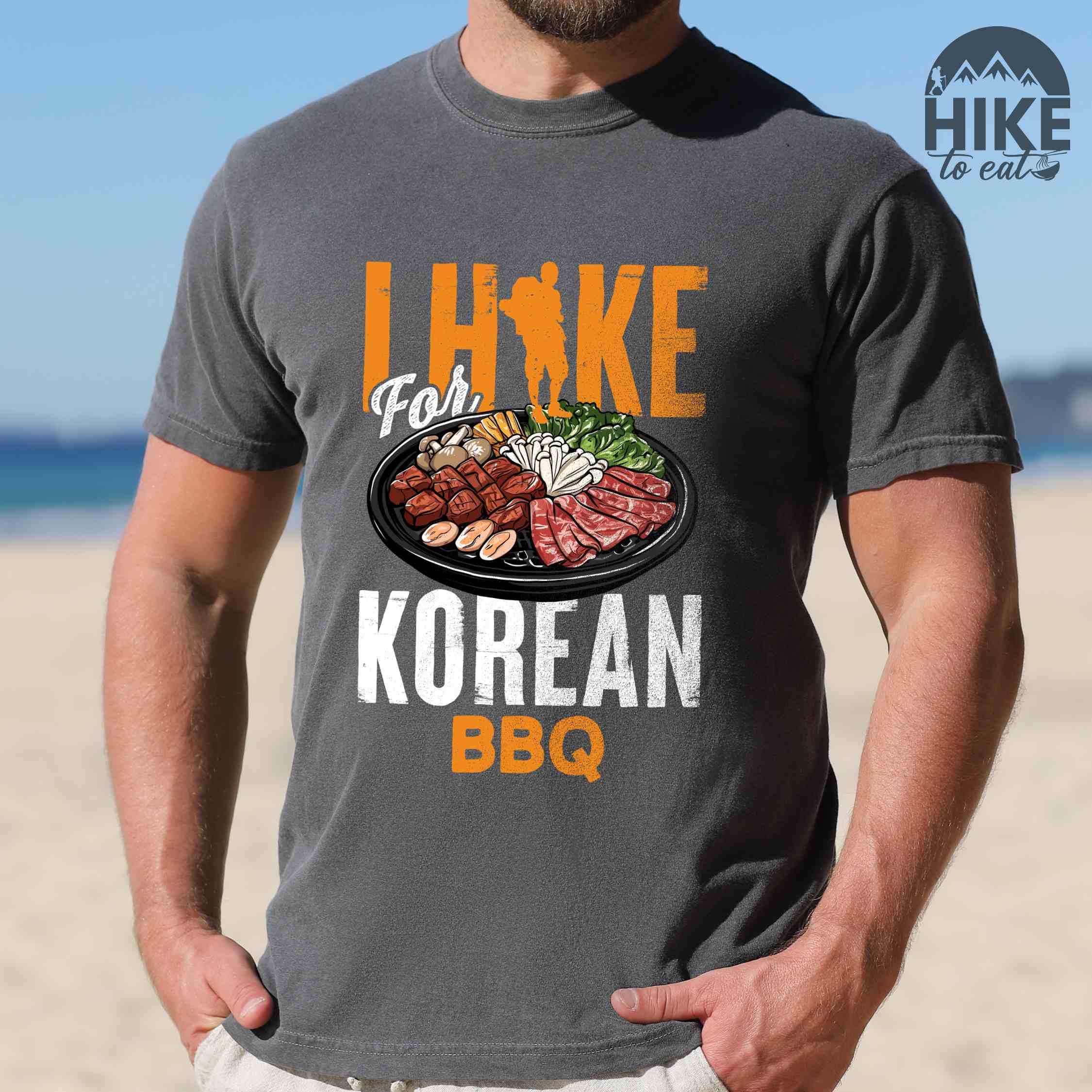 Man wearing pepper "I hike for korean bbq" colored vintage tee with bold typography, illustrated grilling platter korean bbq, mountains, birds, and backpacker for kbbq lovers.