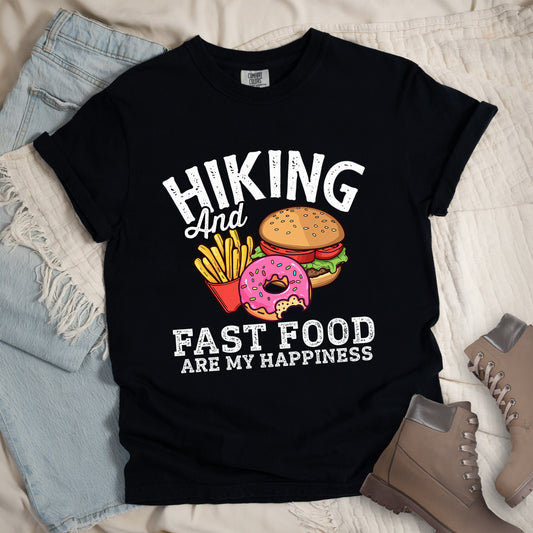 A black t-shirt with white distressed text reading "HIKING And FAST FOOD ARE MY HAPPINESS." The design features colorful illustrations of fast food between the top and bottom text: a hamburger with sesame seed bun, lettuce and tomato, a pink frosted donut with sprinkles (with a bite taken out), and yellow french fries in a red container. The word "And" appears in cursive script. Features a classic crew neck and casual rolled sleeves, styled alongside light wash denim and brown hiking boots. 
