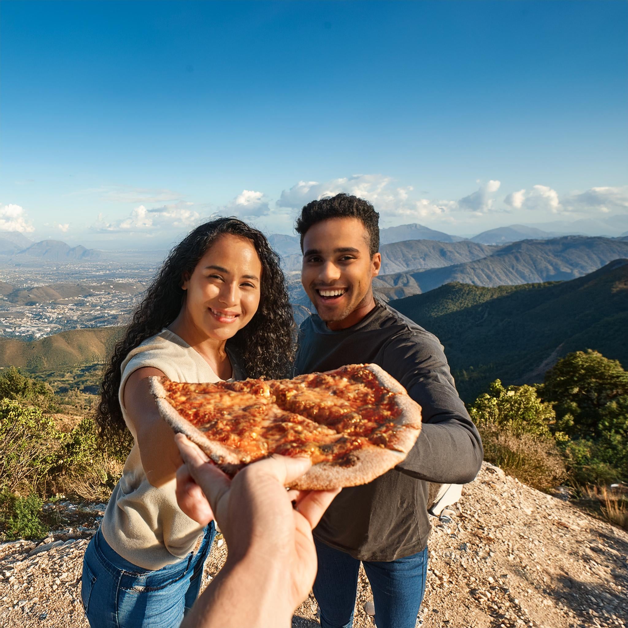 Two smiling hikers on a mountain viewpoint sharing a slice of pizza. A person with curly hair wearing a light sweater and another in a dark long-sleeve shirt hold the pizza together toward the camera. Behind them stretches a panoramic landscape of mountains, valleys, and distant city views under a clear blue sky with scattered clouds.