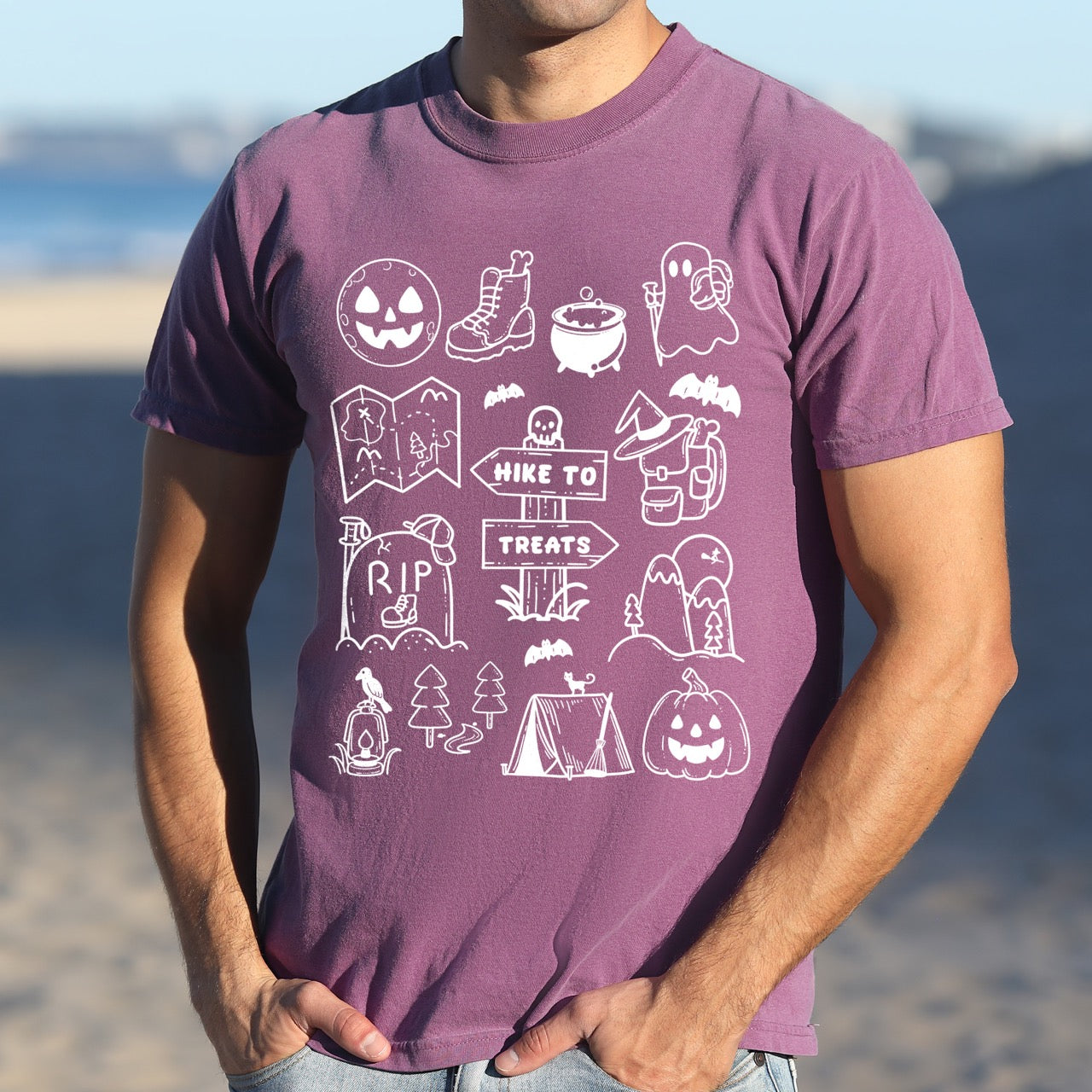 Man wearing Berry (purple) Halloween adventure vintage tee featuring illustrated icon collection with jack-o-lantern boots cauldron ghost backpacker map RIP tombstone Hike to Treats trail sign tent pumpkin bats and trees on Comfort Colors shirt for spooky season celebrations for hikers, foodies and adventurers