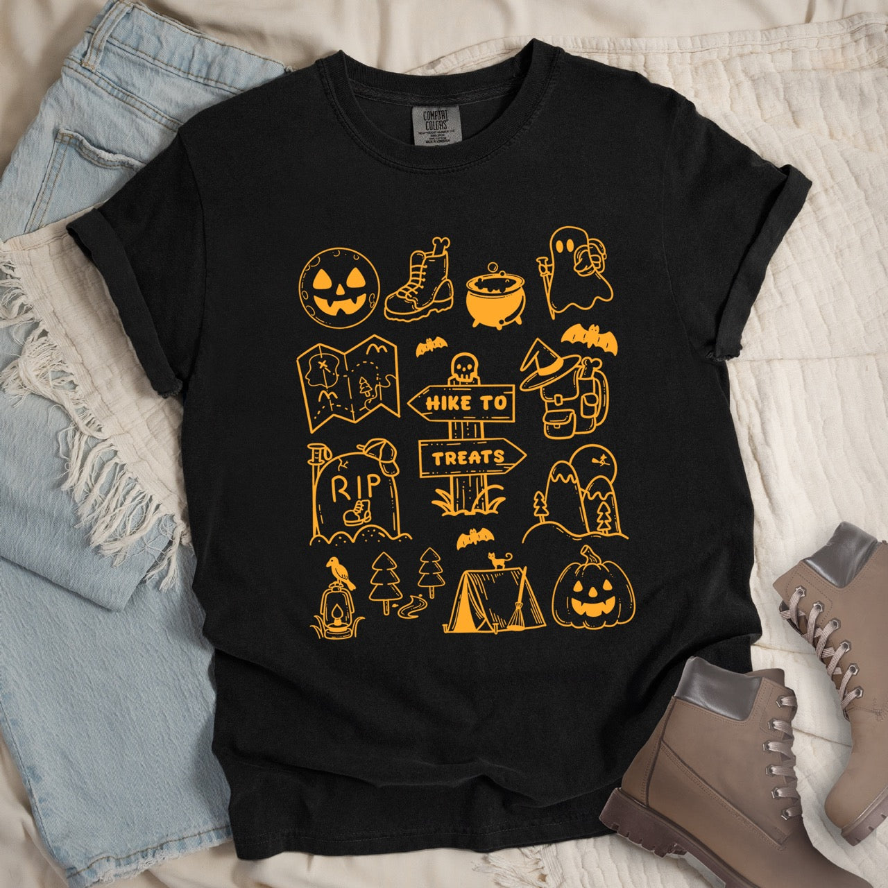 Black Halloween adventure vintage tee featuring illustrated icon collection with jack-o-lantern boots cauldron ghost backpacker map RIP tombstone Hike to Treats trail sign tent pumpkin bats and trees on Comfort Colors shirt for spooky season celebrations for hikers, foodies and adventurers - laid flat with jeans and boots