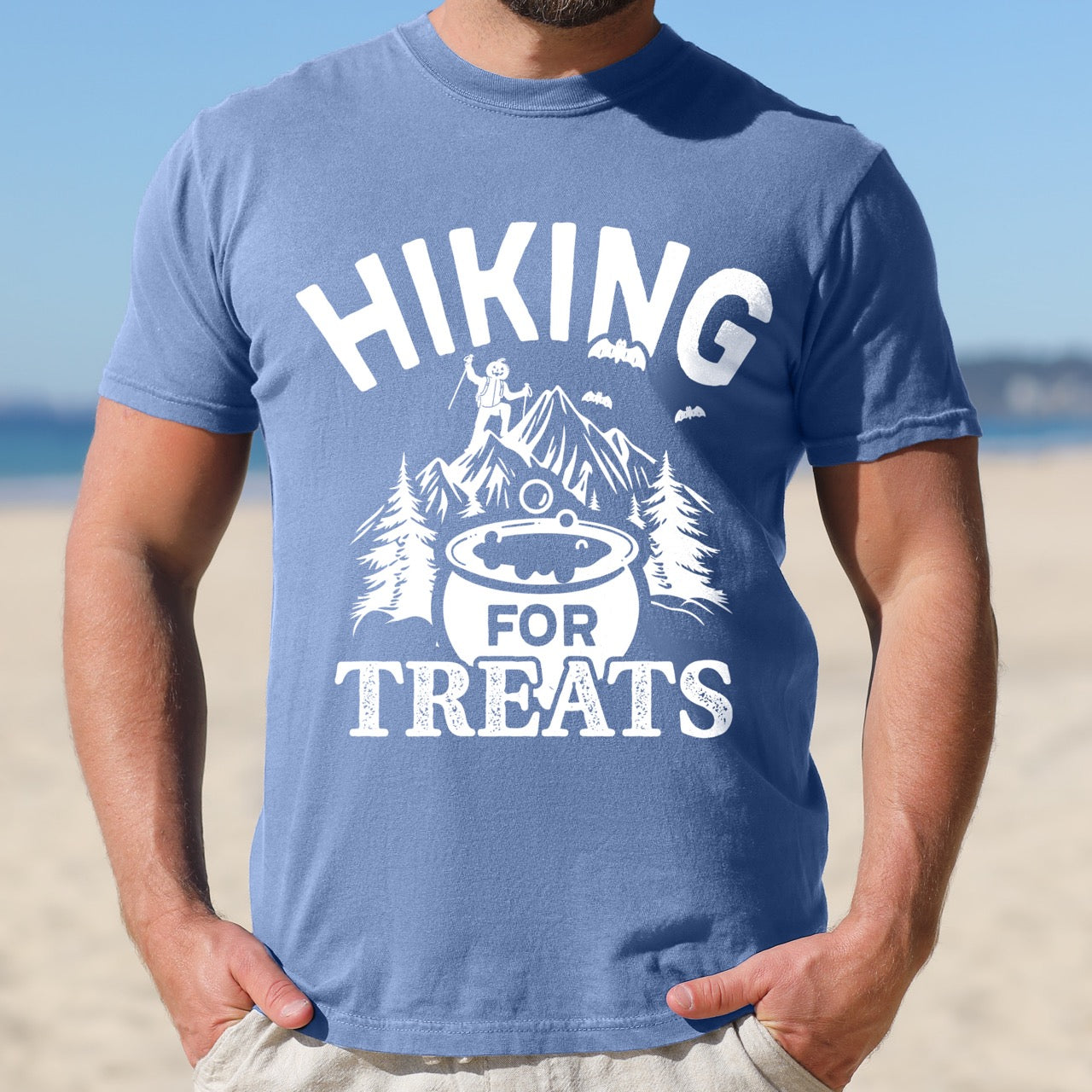 Man wearing blue jean Halloween vintage tee Hiking for Treats featuring curved typography bubbling cauldron with candy mountain landscape with character on peak pine trees and flying bats on Comfort Colors shirt for spooky season celebrations for hikers, foodies and adventurers. 