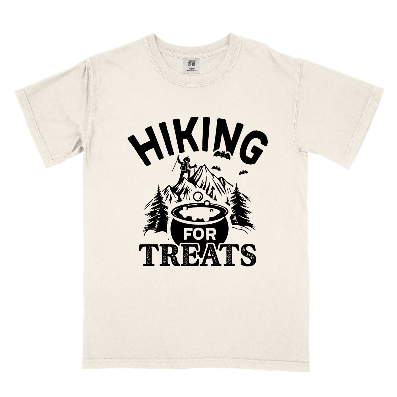 Ivory  Halloween vintage tee Hiking for Treats featuring curved typography bubbling cauldron with candy mountain landscape with character on peak pine trees and flying bats on Comfort Colors shirt for spooky season celebrations for hikers, foodies and adventurers. 